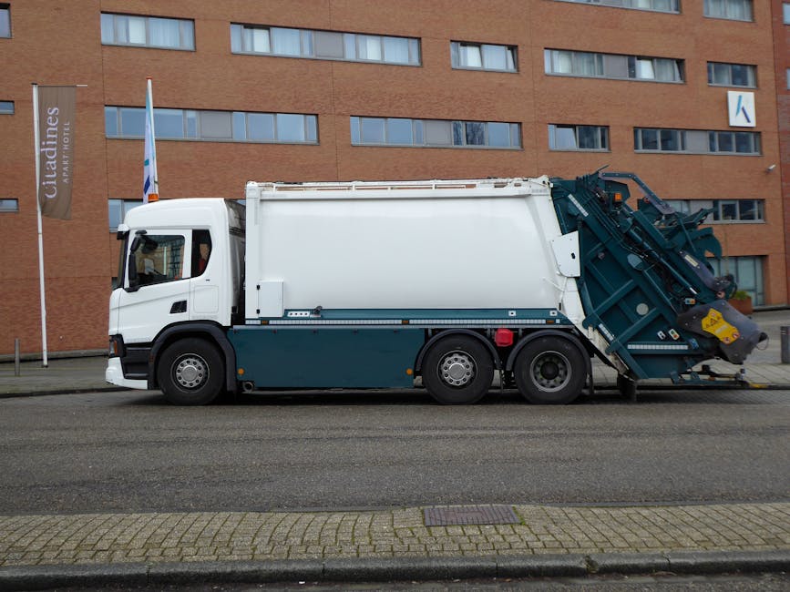 A group of sanitation workers wearing orange uniforms and black helmets are actively collecting waste from a large, open-topped red garbage truck parked on a city street. The truck features a green an