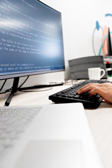 A young man with dark, wavy hair is seated at a desk in a professional office setting, working intently on a desktop computer. The monitor displays lines of code or text, suggesting programming or dat