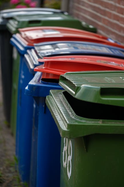 A row of five large wheelie bins placed along a pavement beside a brick wall, each with a different coloured lid—green, blue, red, and a darker green—organized in sequence. The green bin is closest to
