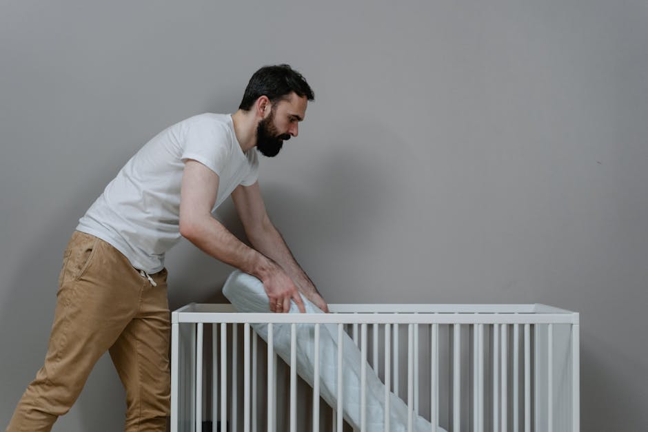 A man with a dark beard, wearing a white T-shirt and tan pants, is seen leaning over a white vertical slatted crib or bed frame against a plain gray wall. He appears to be lifting or adjusting a mattr